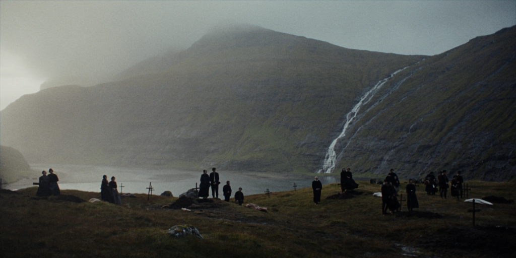 Villagers gather near graves beneath a mountain and waterfall in No Rest for the Wicked