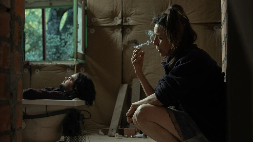 A woman smokes while a man lies on a bench in an unfinished room in La Bola Negra