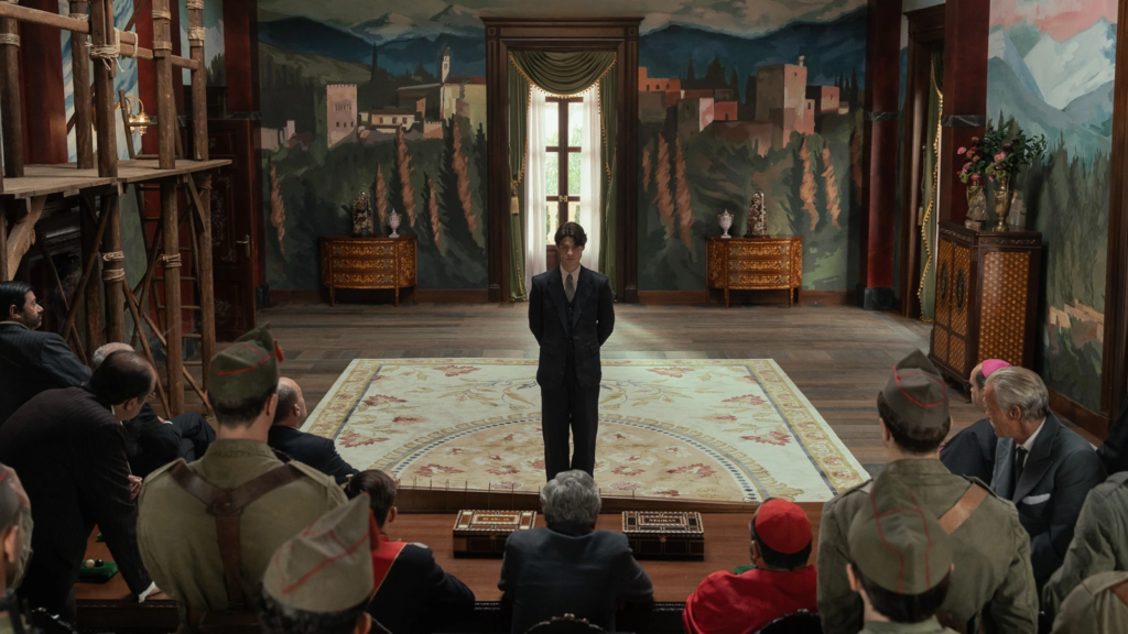 Carlos stands alone before a group of men in a formal room in La Bola Negra