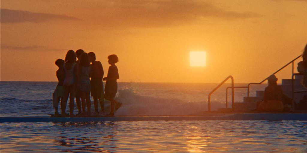 A group of people gather beside a swimming pool at sunset, silhouetted against a glowing square sun in Everytime.