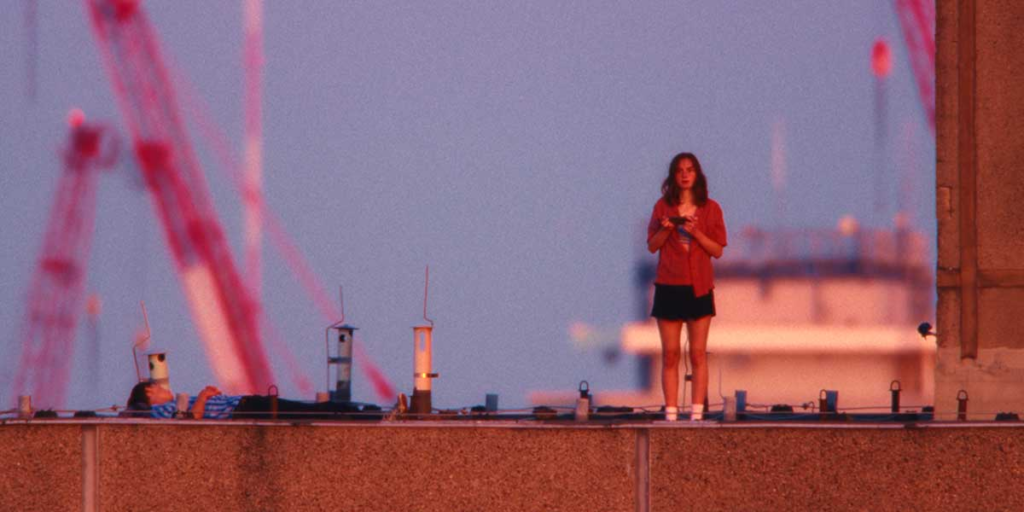 A young woman stands on a rooftop at dusk while another person lies nearby, framed against a pink and blue sky in Everytime.