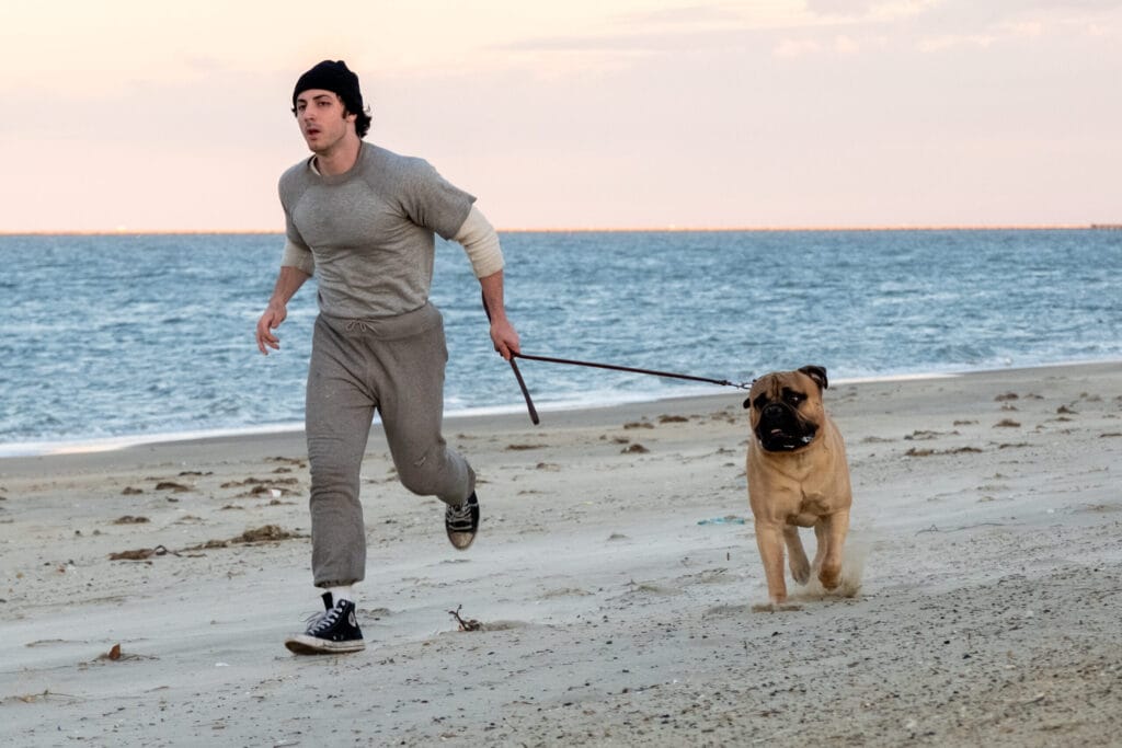 Anthony Ippolito as Sylvester Stallone runs with a dog along a beach in I Play Rocky