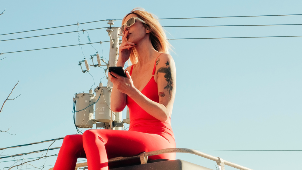 Woman in a bright red outfit sits outdoors beneath power lines in The Snake